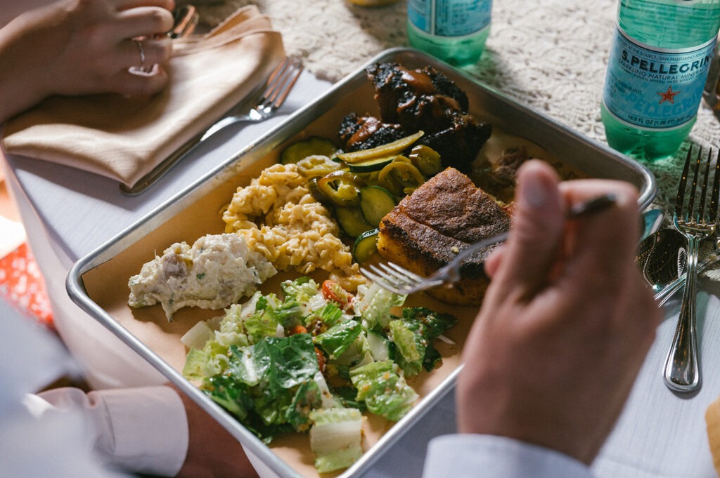 A guest digs into a metal tray of brisket, mac and cheese, and cornbread