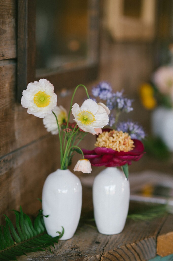 White ceramic vases hold poppies and a deep red peony on a rustic wood table