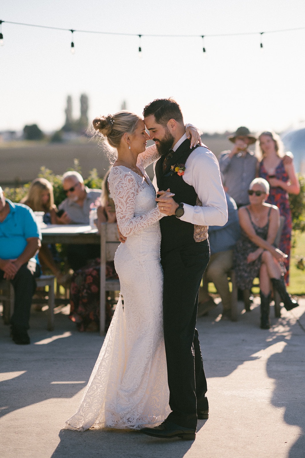 Bride and groom sway together outdoors during their first dance as guests watch 