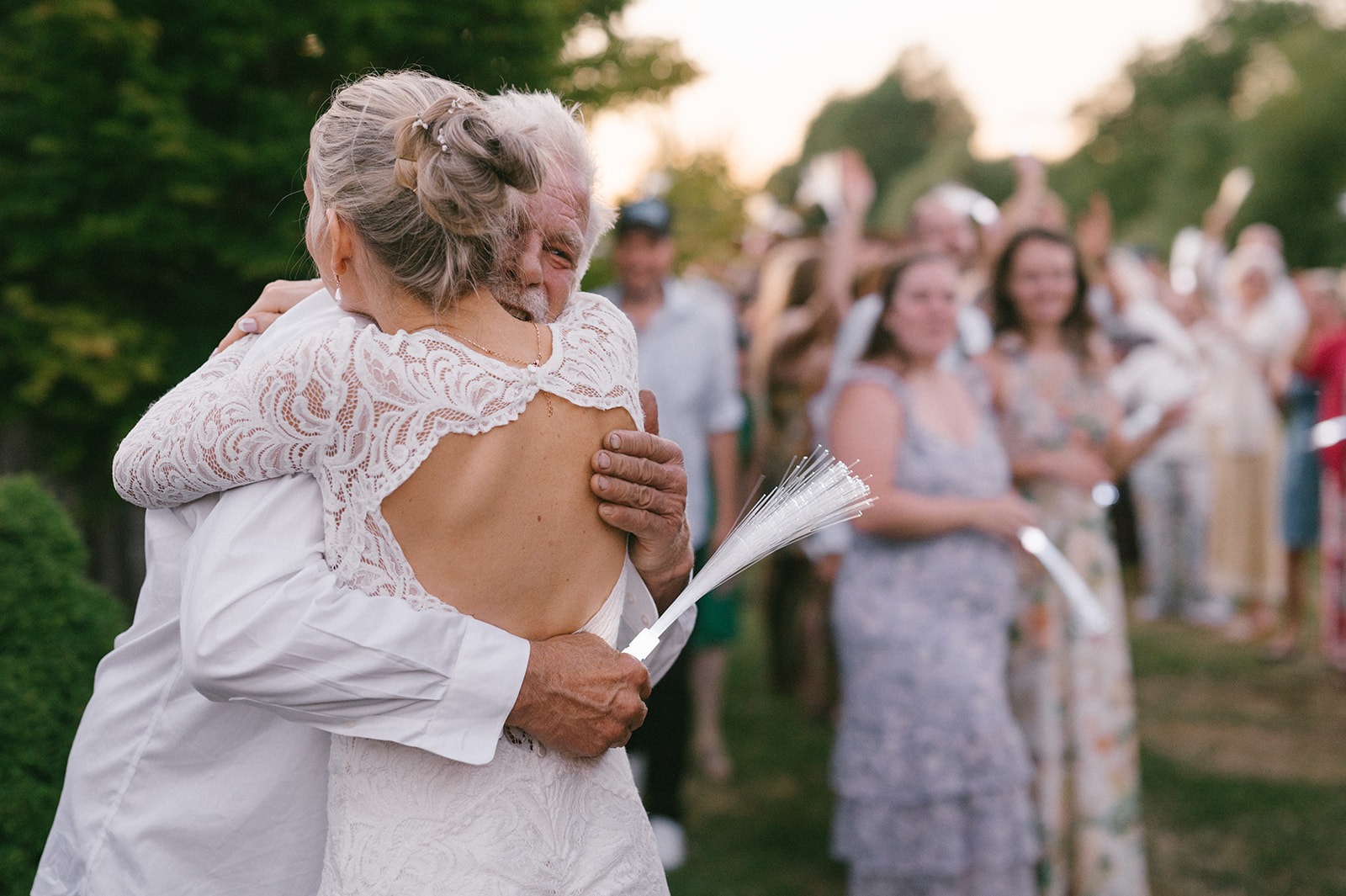 Bride embraces her father as guests cheer with ribbon wands behind them