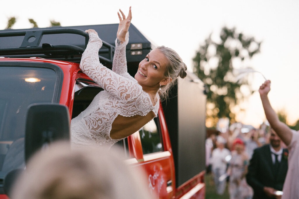 Bride leans out of the truck window, arm raised, as guests cheer her on