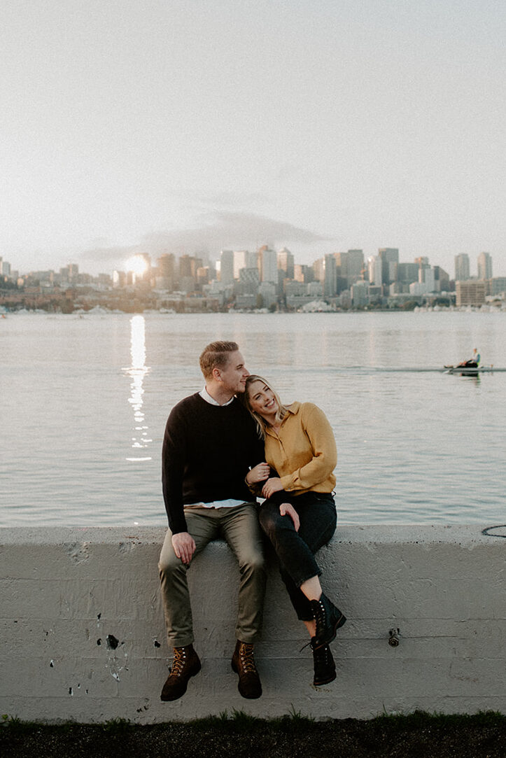 Couple sitting along the Lake Union waterfront with the Seattle skyline during a Gas Works Park engagement session