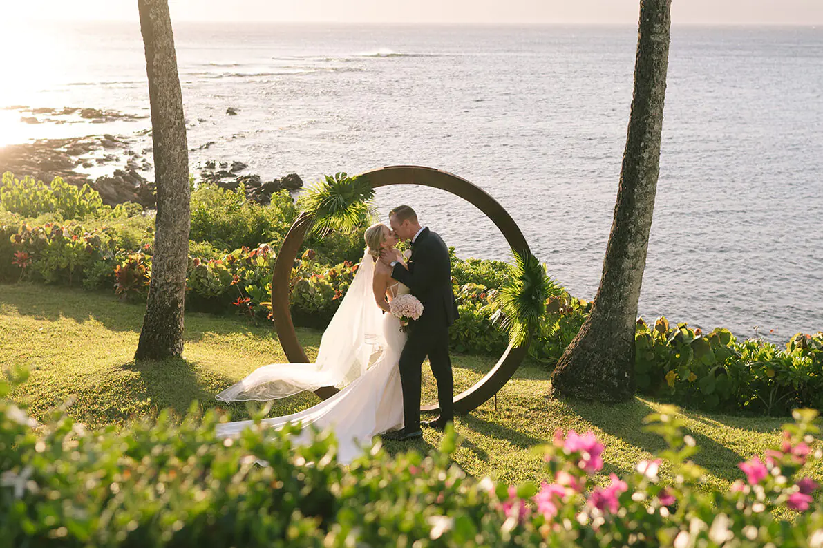 Bride and groom sharing their first kiss beneath a circular arch overlooking the ocean at Merriman’s Kapalua during golden hour.