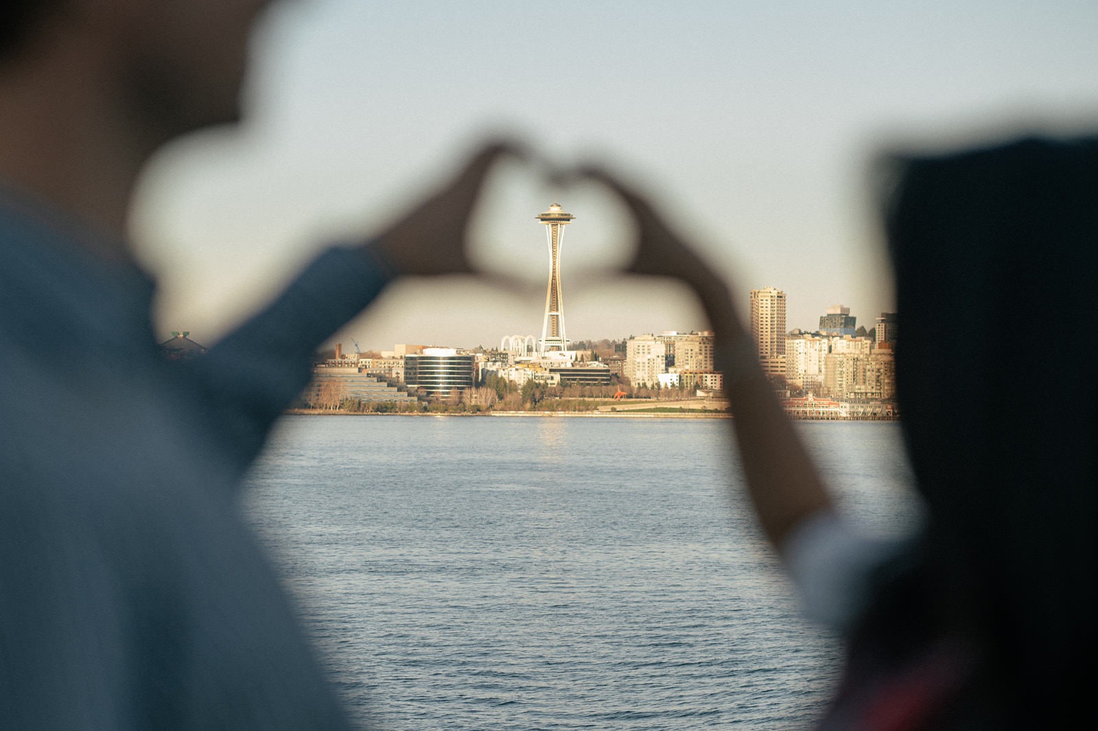 Couple making a heart shape with their hands around the Space Needle across the water