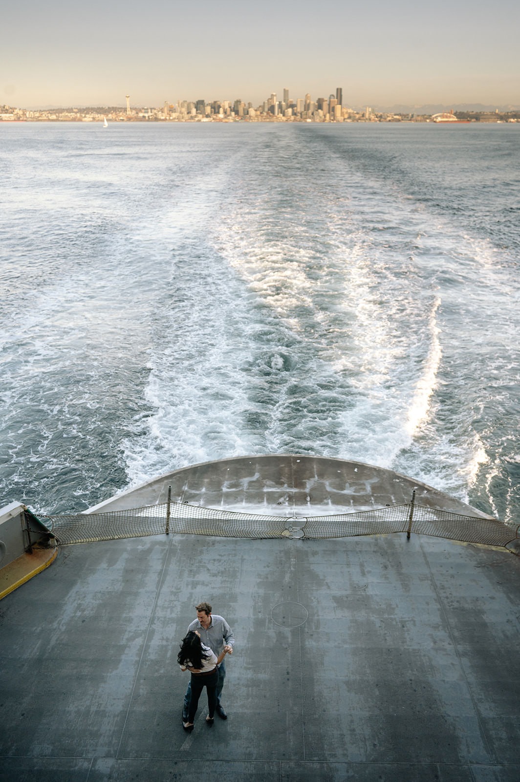 View from the back of a Washington State Ferry with wake lines, Seattle skyline, and a couple standing on the lower deck