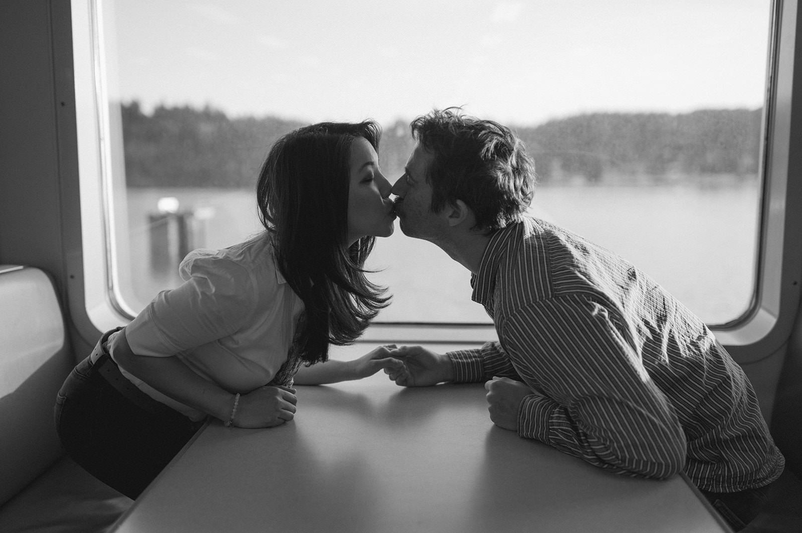 Black and white photo of a couple kissing across a ferry table by the window