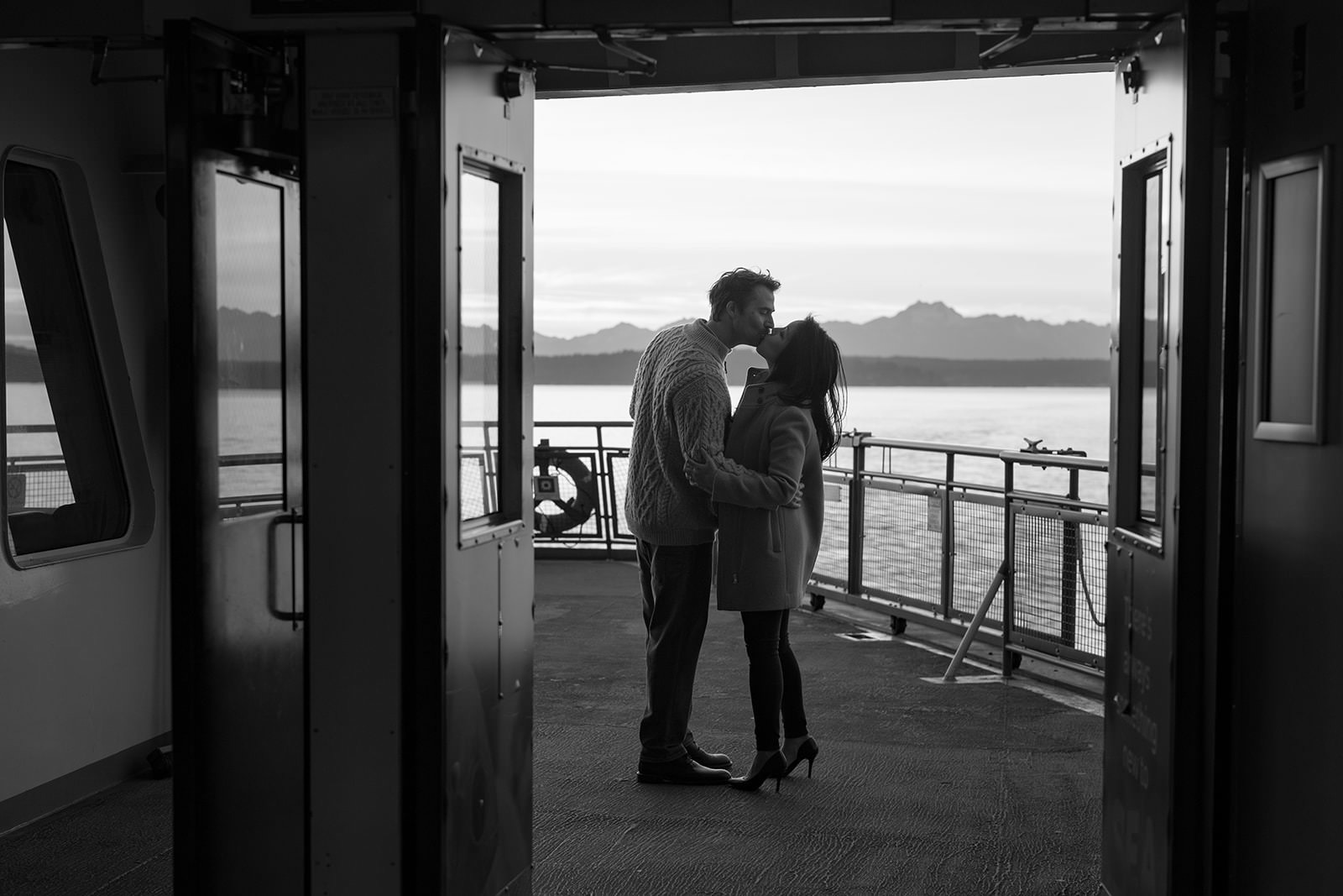 Black and white photo of a couple kissing in the ferry doorway with water and distant mountains behind them