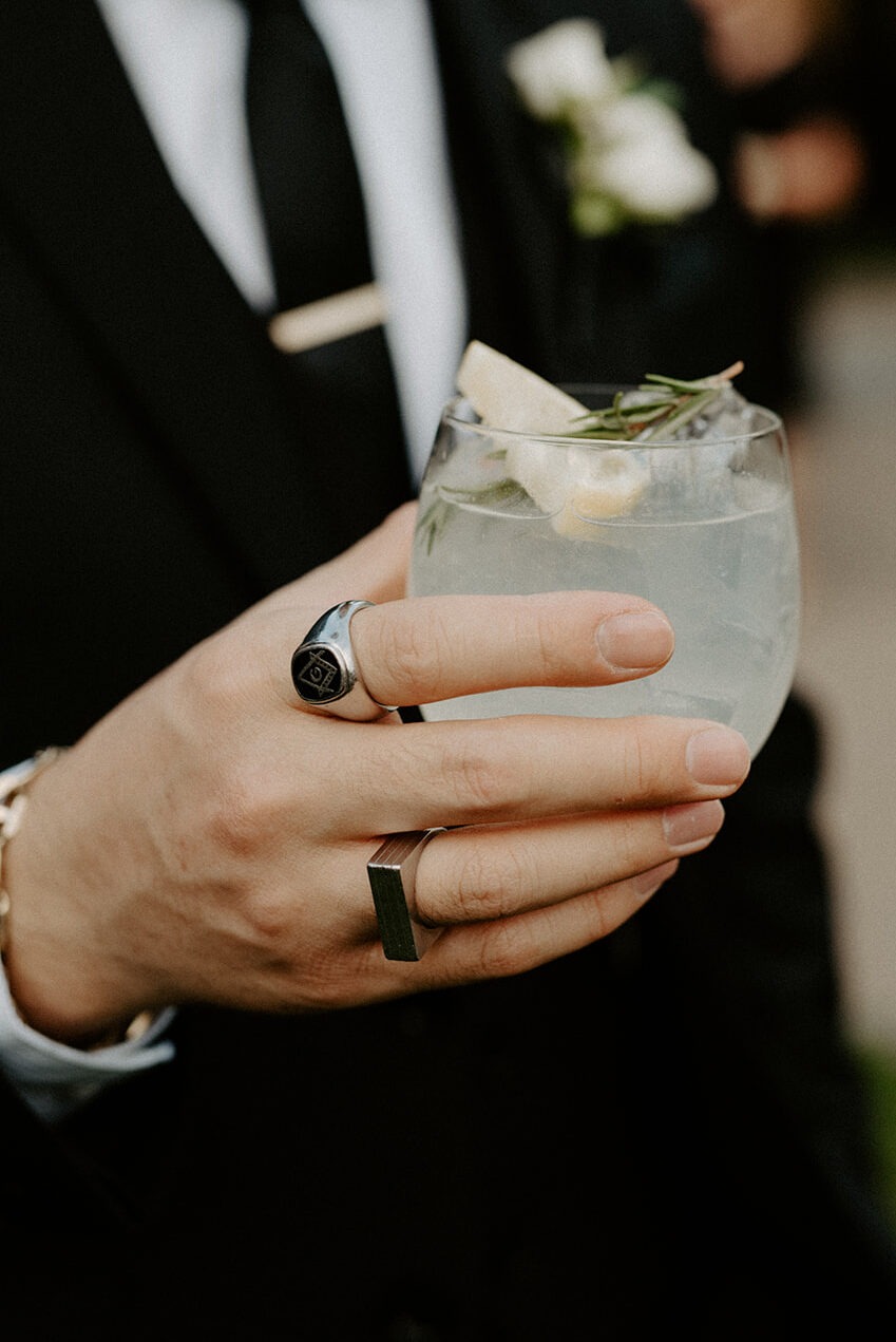 Close detail of the groom's hand holding a cocktail glass, his wedding ring and white boutonniere visible