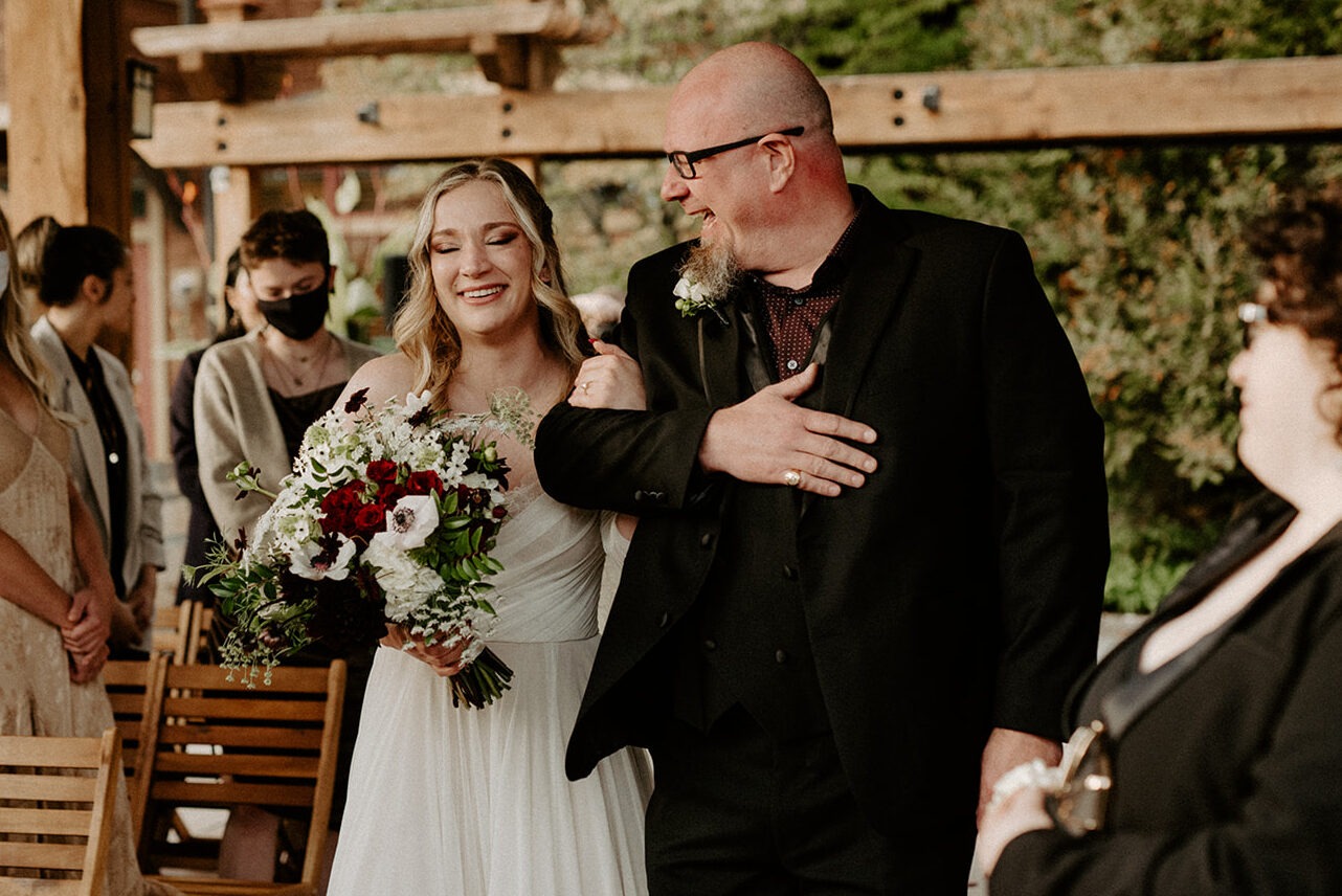 Bride walking down the aisle arm in arm with her father, smiling joyfully, holding a bouquet of dark red and white flowers at Willows Lodge