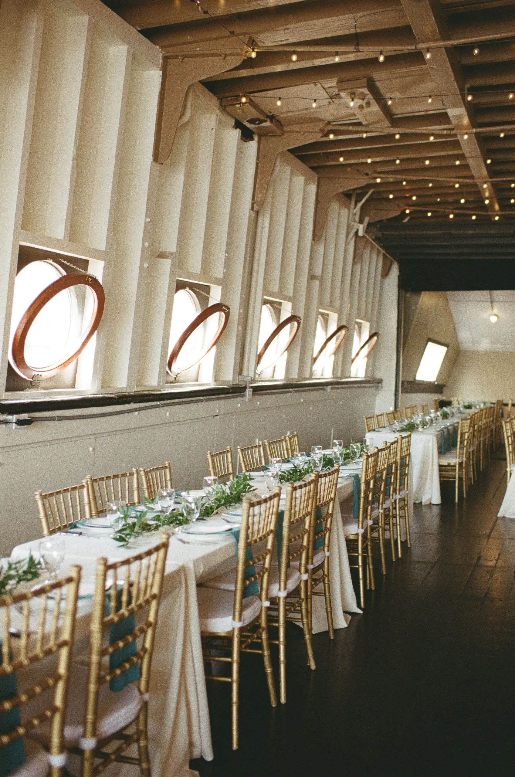 MV Skansonia wedding reception room with long tables, gold chiavari chairs, greenery runners, and porthole windows lined with string lights