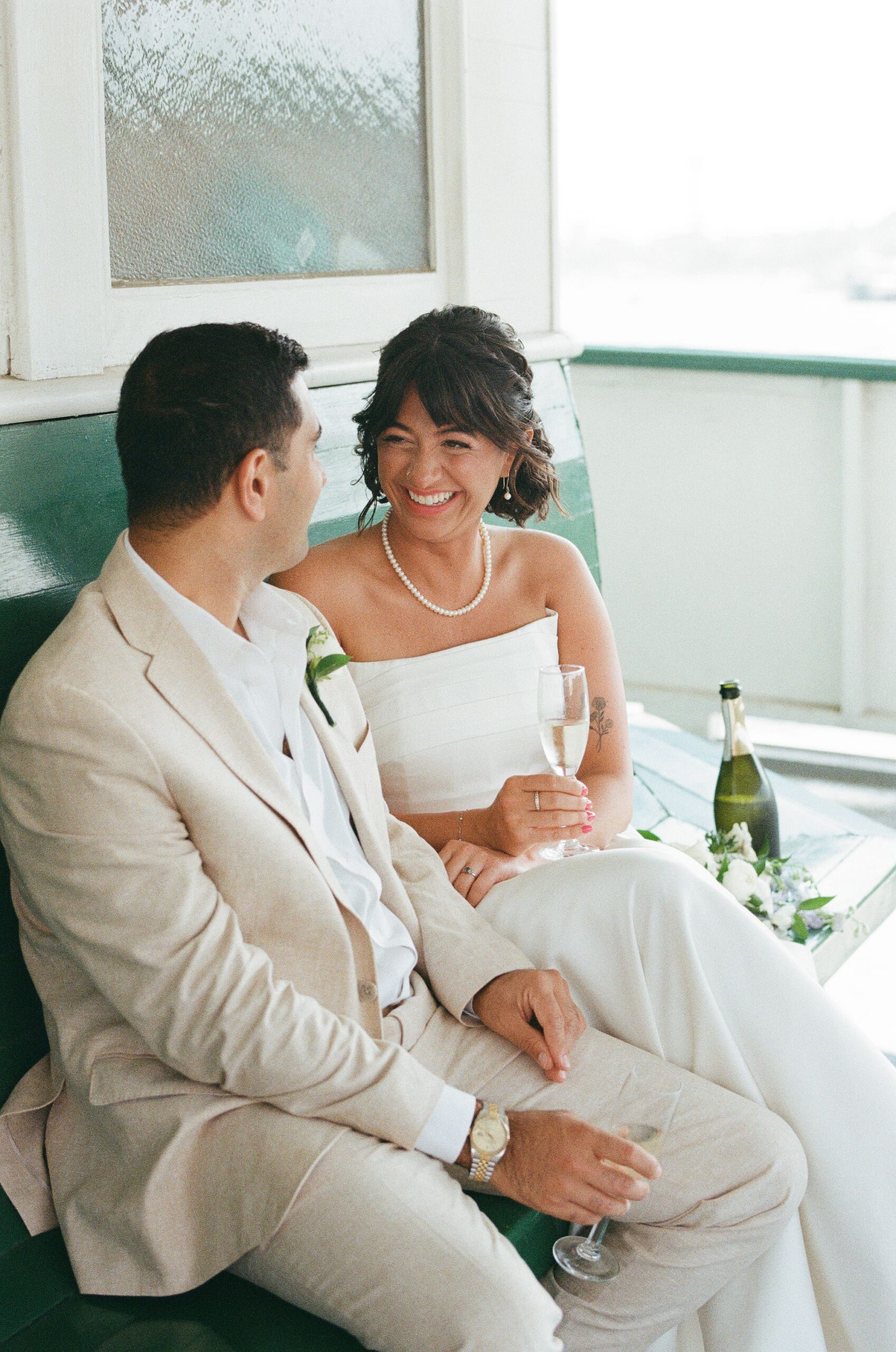 Bride and groom laughing together on the original green ferry benches inside MV Skansonia, champagne in hand