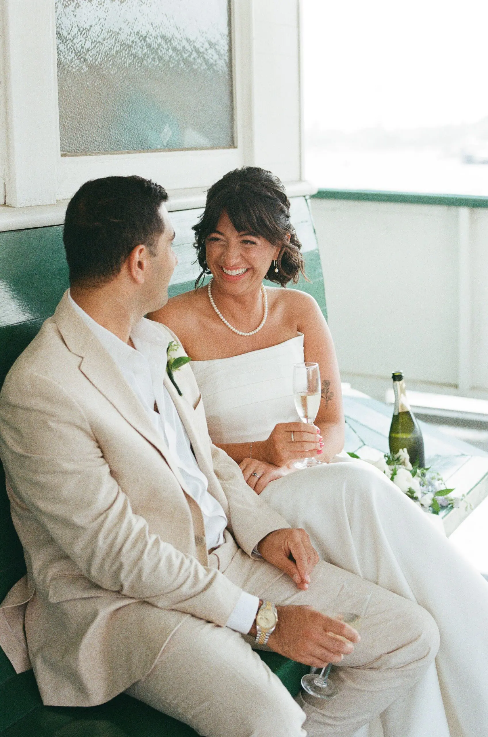 Bride and groom laughing together on the original green ferry benches inside MV Skansonia, champagne in hand