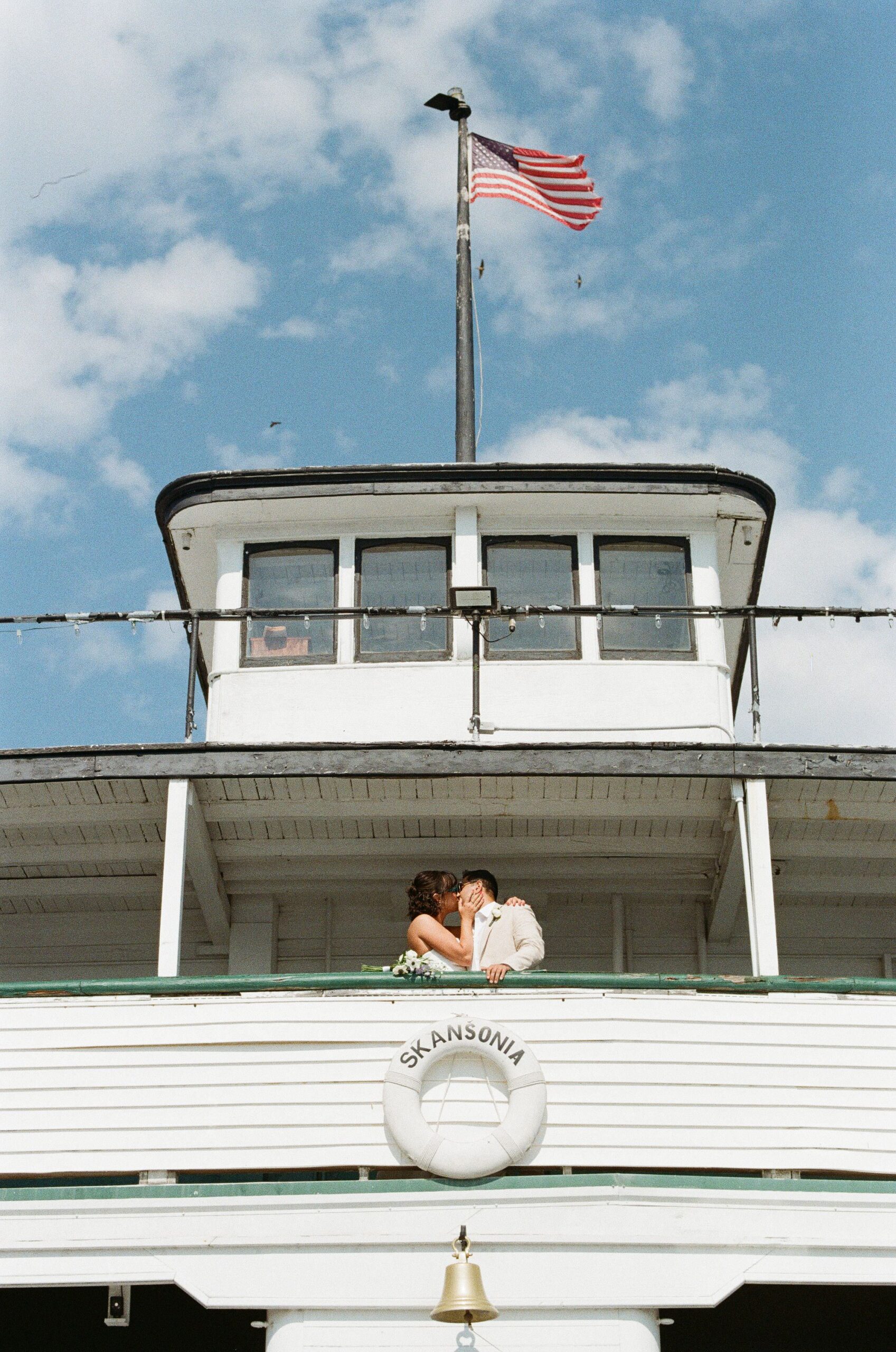 Bride and groom kissing on the upper deck of MV Skansonia with an American flag and blue sky above