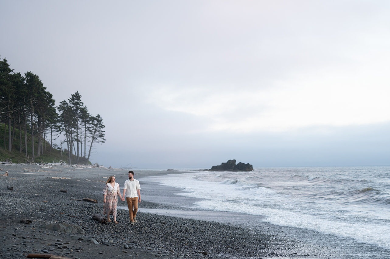 Couple walking hand in hand along Ruby Beach at dusk, waves crashing on the dark pebble shoreline, Olympic National Park Washington