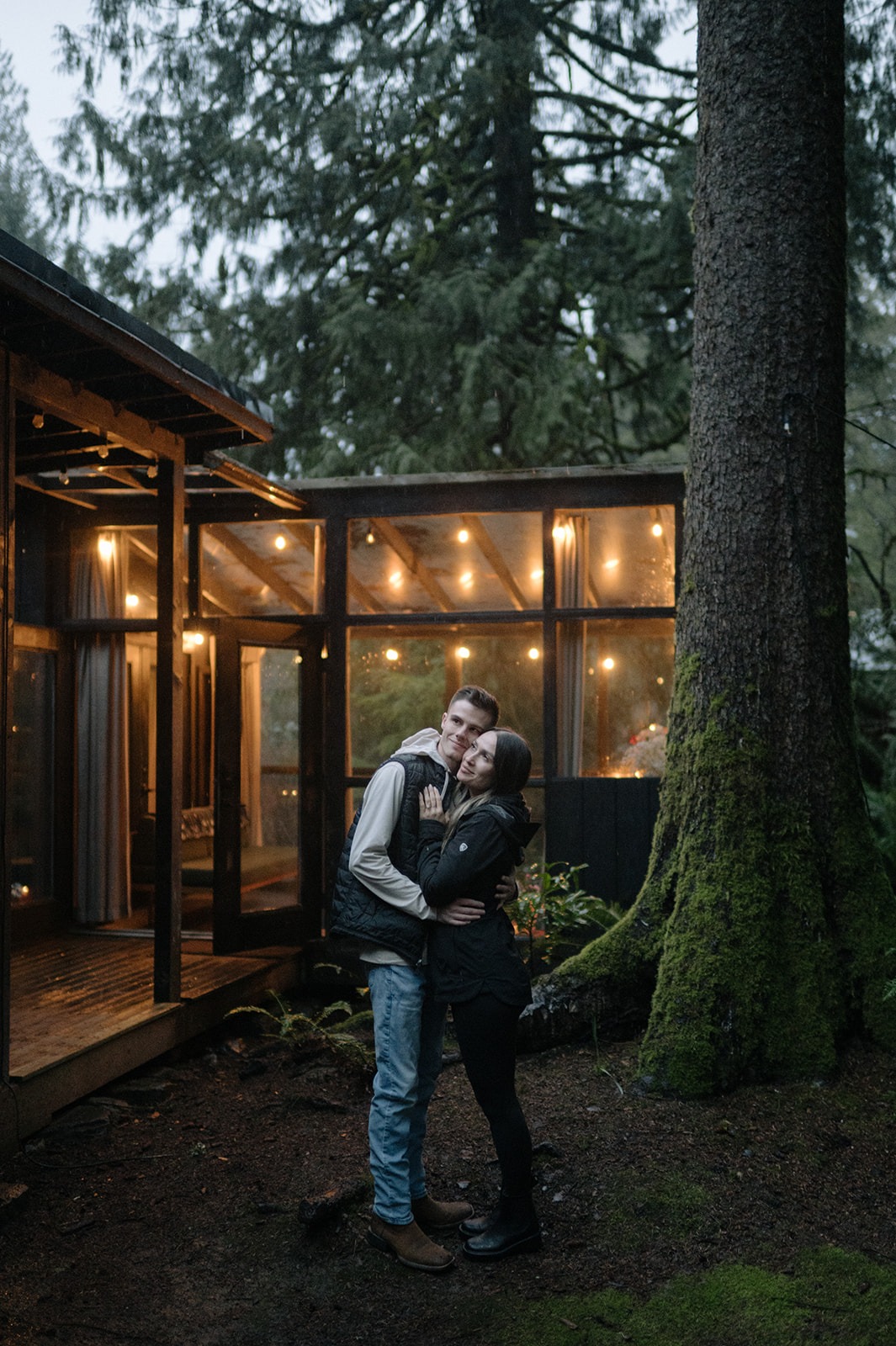 Newly engaged couple embraces outside their cabin beside a moss-covered old-growth tree at Canyon Creek Cabins

