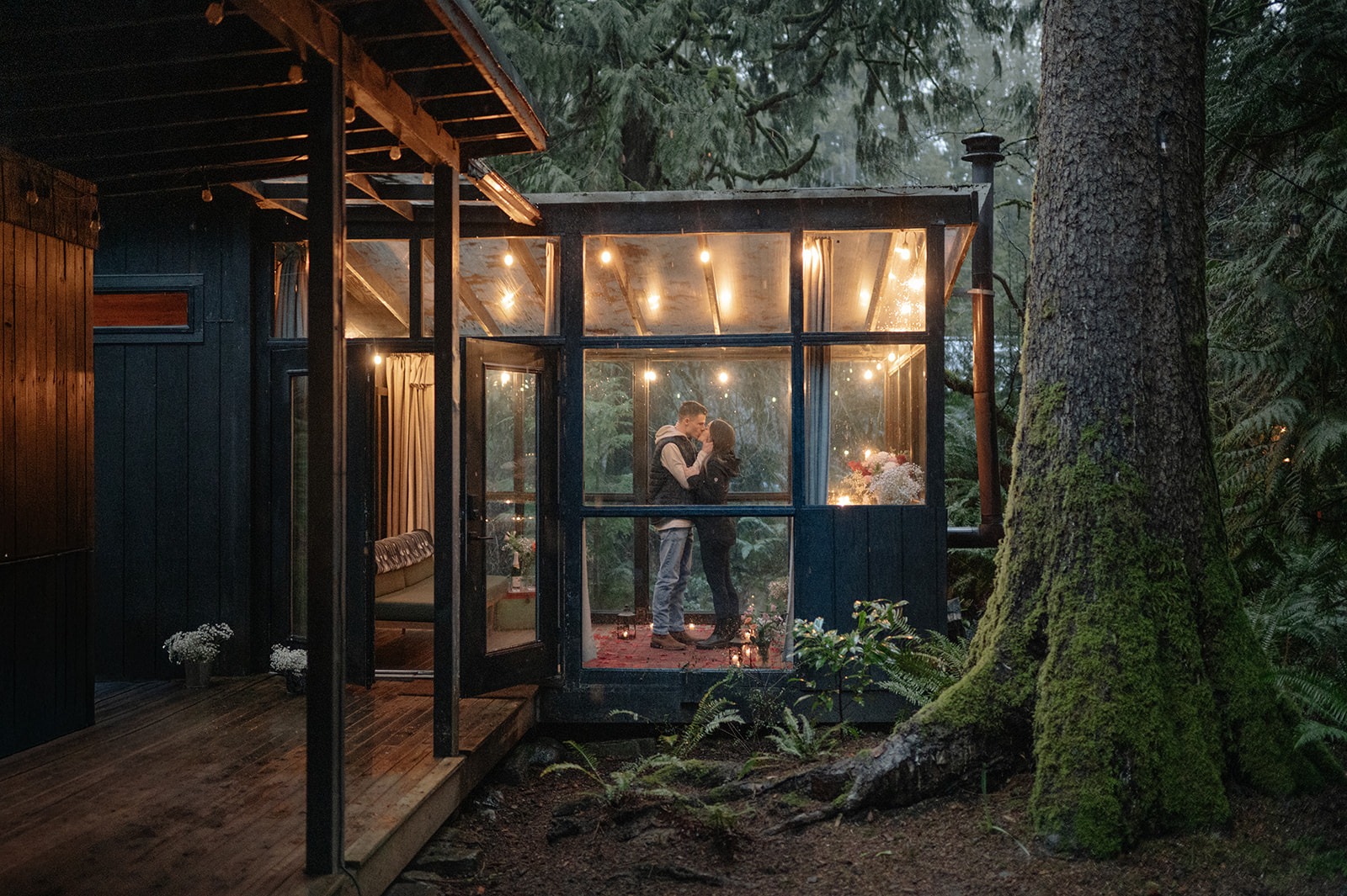 Wide view of the glass sunroom at Canyon Creek Cabins with the newly engaged couple embracing inside, surrounded by the Cascade rainforest