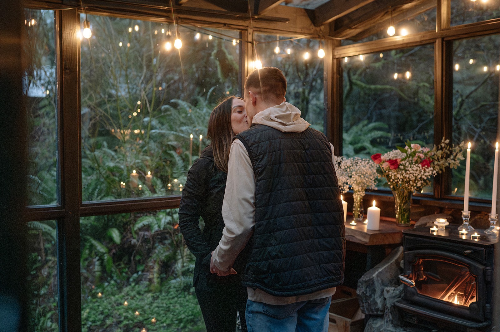 Newly engaged couple shares a kiss beside the wood stove inside the glass sunroom at Canyon Creek Cabins