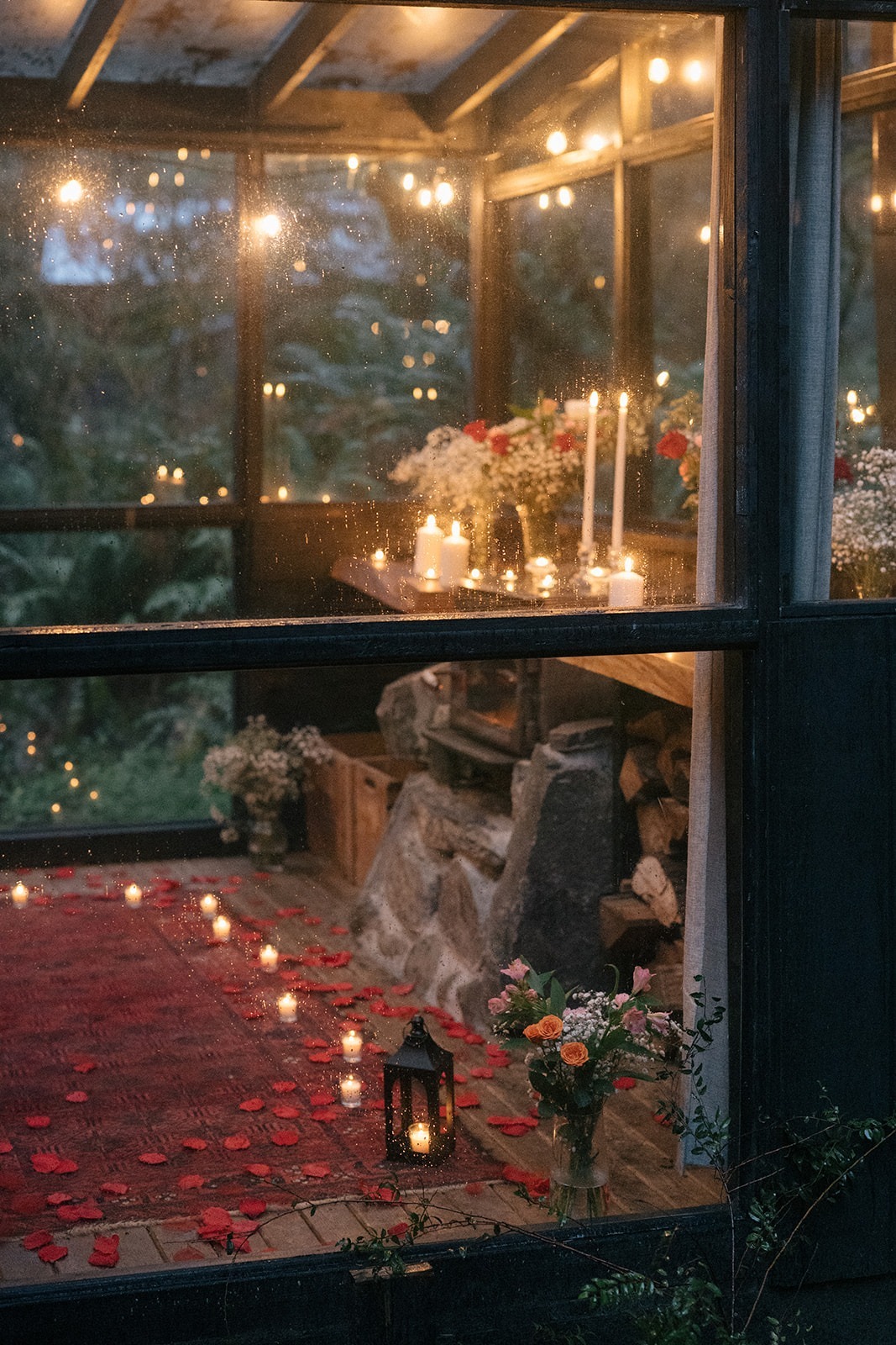 Candlelit proposal setup with red rose petals and florals seen through the rain-speckled glass walls of a Canyon Creek Cabins sunroom