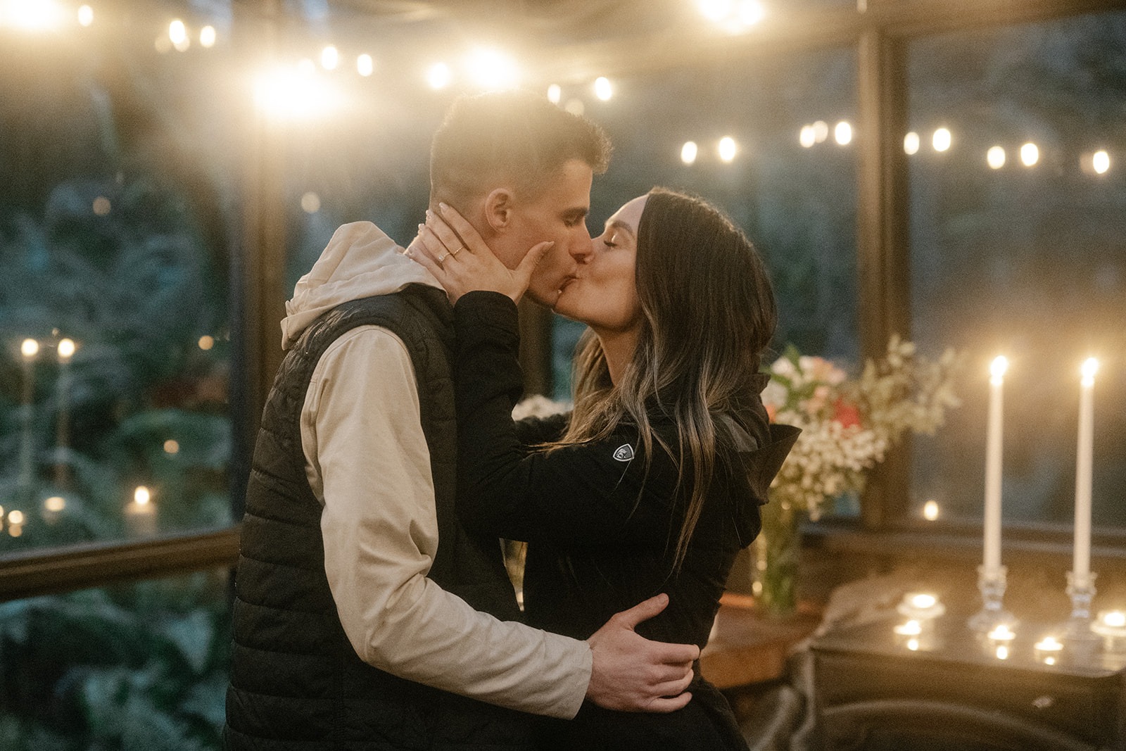 Close romantic kiss between newly engaged couple with taper candles glowing beside them inside a PNW cabin