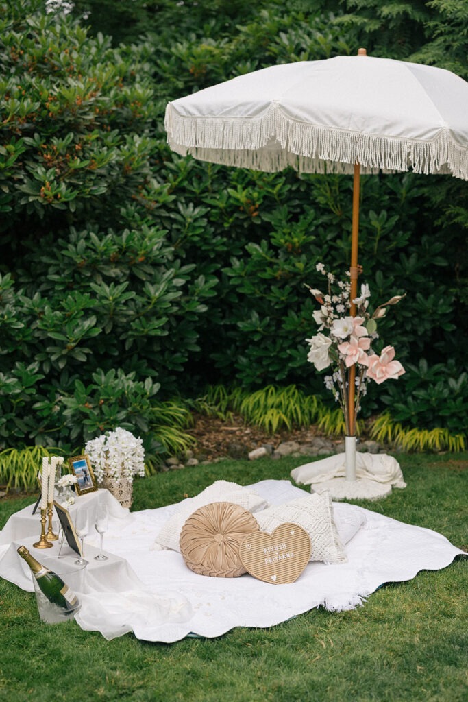 Boho picnic setup with fringed umbrella, champagne, and framed photos styled by Picnic Party Seattle for a surprise proposal