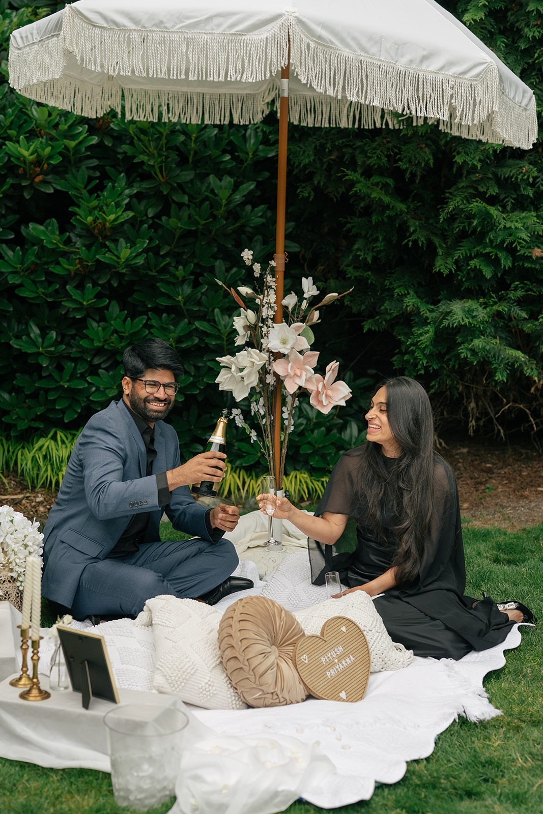 Newly engaged Piyush and Priyanka toast on their picnic blanket under a fringed umbrella at Willows Lodge in Woodinville