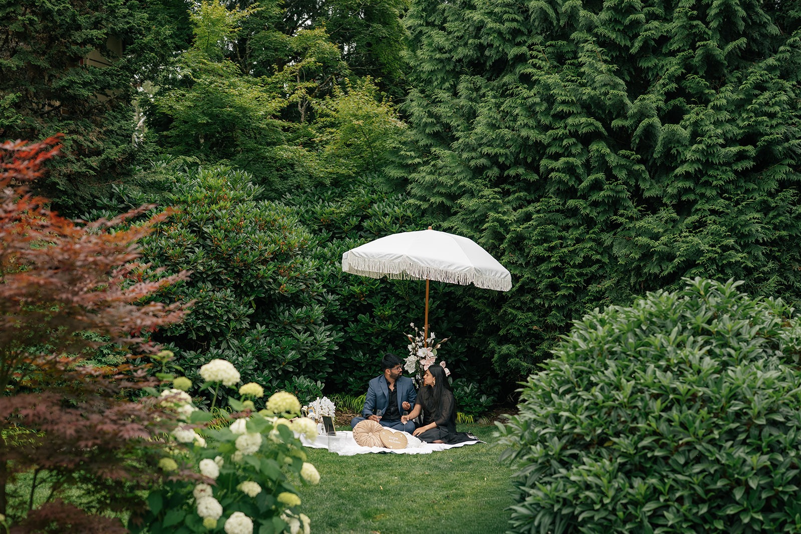 Piyush and Priyanka relax at their picnic setup under a white fringed umbrella, tucked into the lush gardens of Willows Lodge in Woodinville, WA