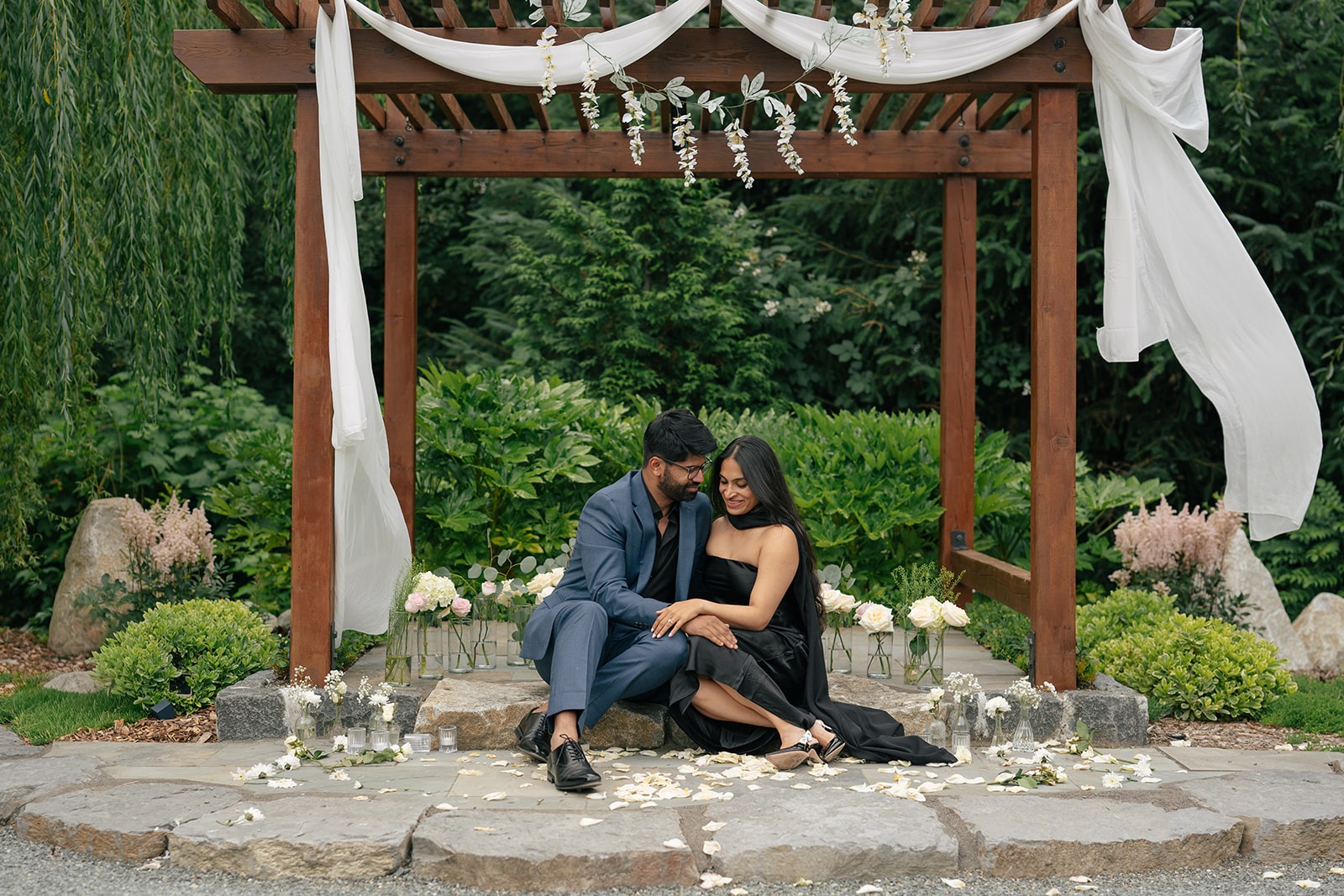 Piyush and Priyanka cuddle on the stone steps beneath their floral proposal arch at Willows Lodge in Woodinville