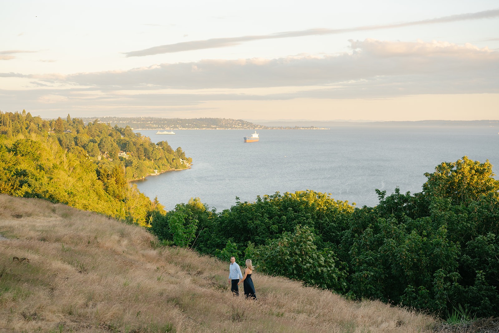 Couple stands on a grassy hillside overlooking Puget Sound as a cargo ship drifts by