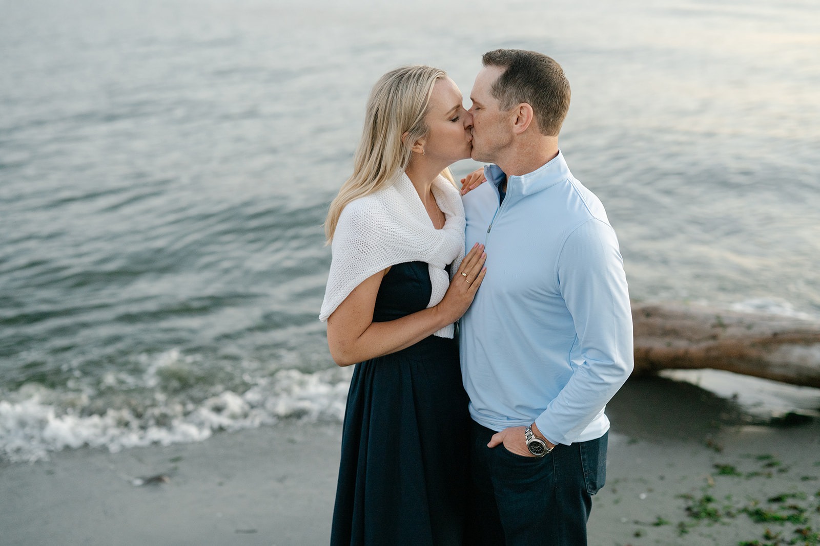 Couple shares a kiss by the water at sunset, one wrapped in a white knit shawl