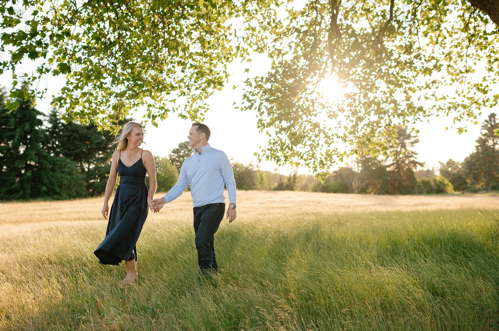 Couple walks hand in hand through a golden grass meadow beneath a tree canopy at sunset
