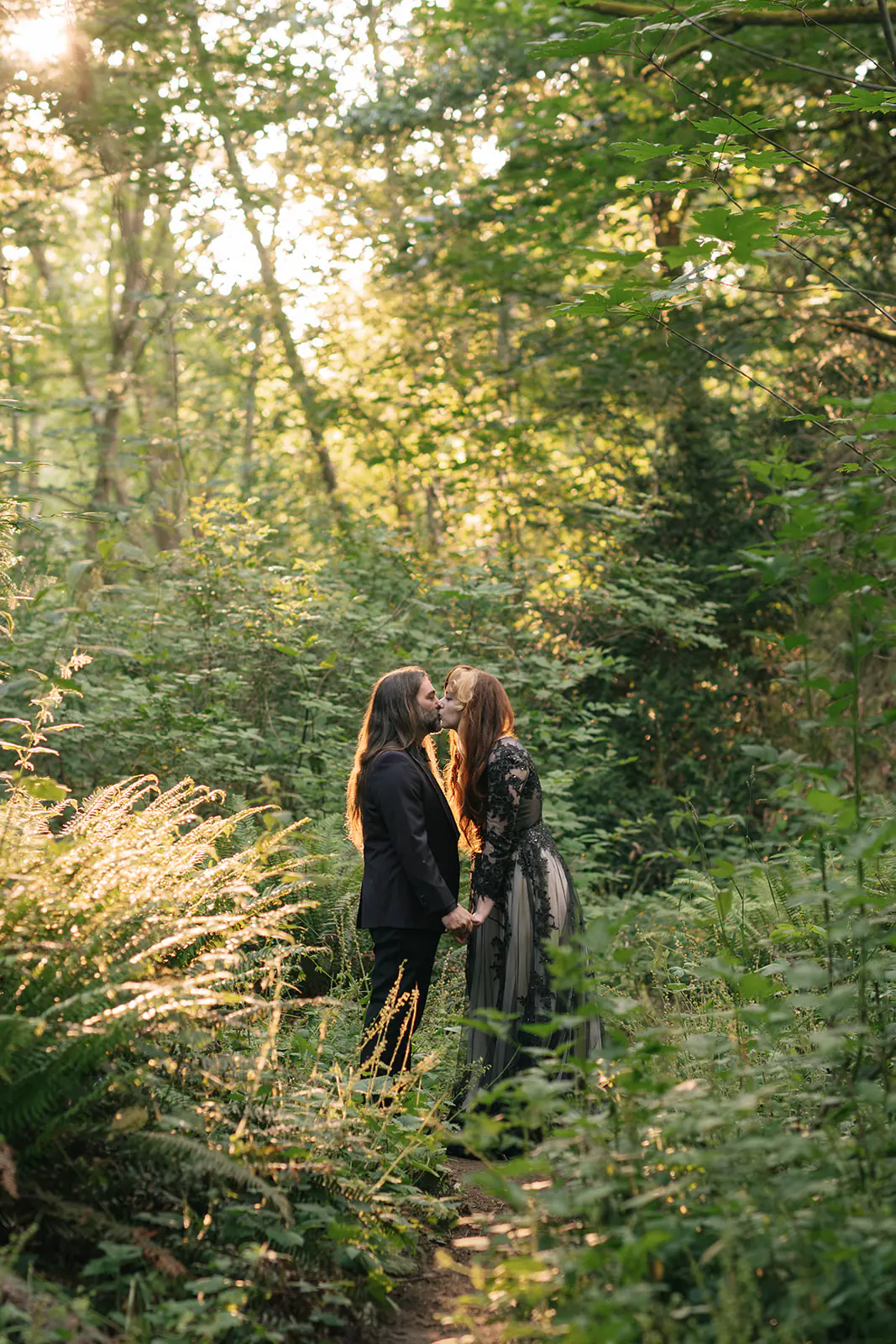 Couple kisses on a sunlit forest path, warm light glowing through the canopy behind them