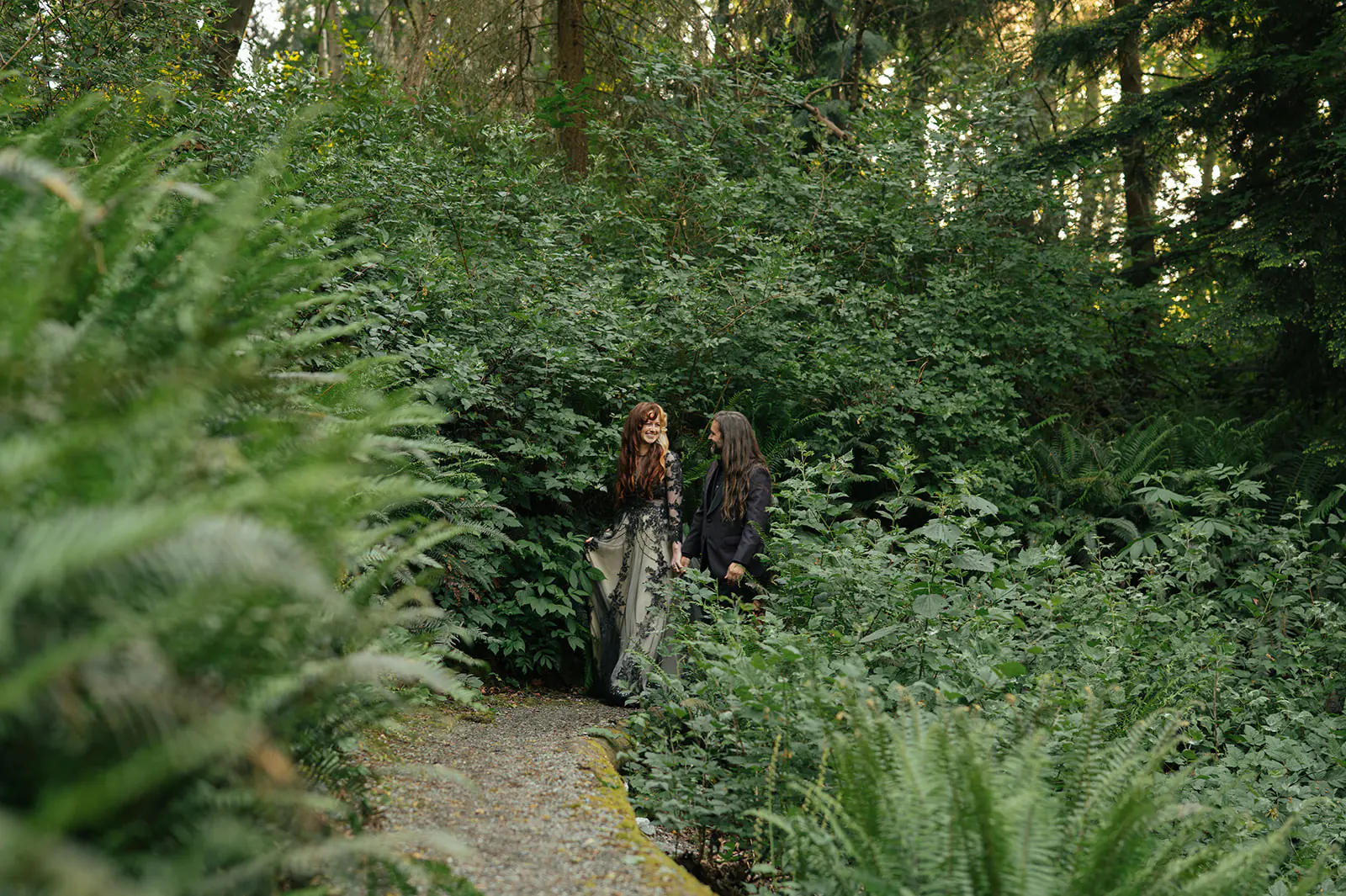 Couple walks a gravel path through a fern-filled forest, seen from a distance