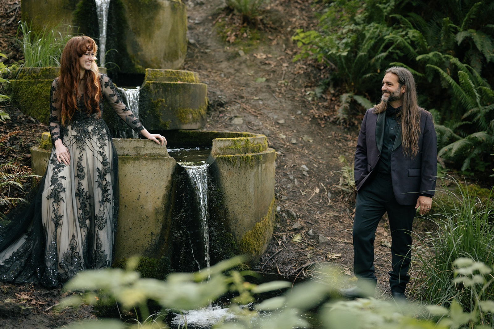Bride smiles beside the waterfall, her gown catching the light on the mossy stone