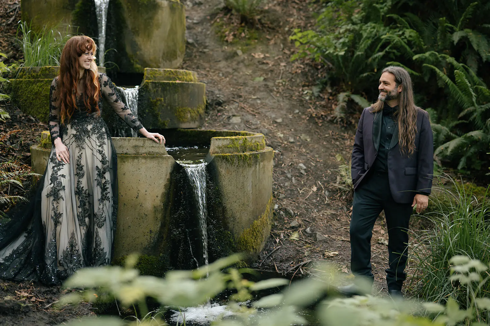 Bride smiles beside the waterfall, her gown catching the light on the mossy stone