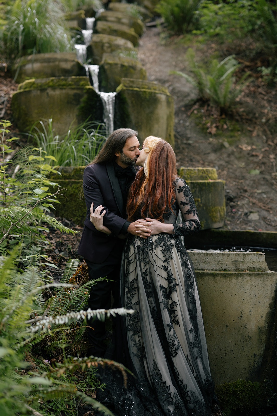 Couple kisses beside the cascading stone waterfall, moss covering the terraces around them