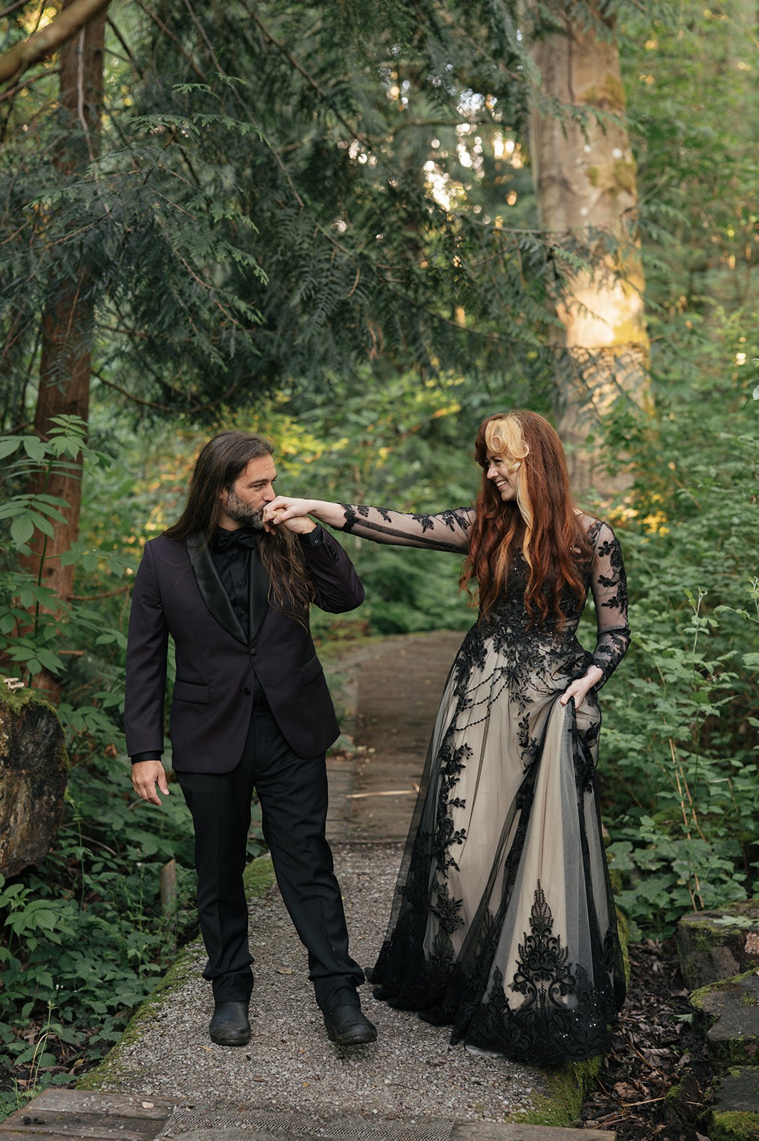 Groom kisses the bride's hand as they pause together on the forest path
