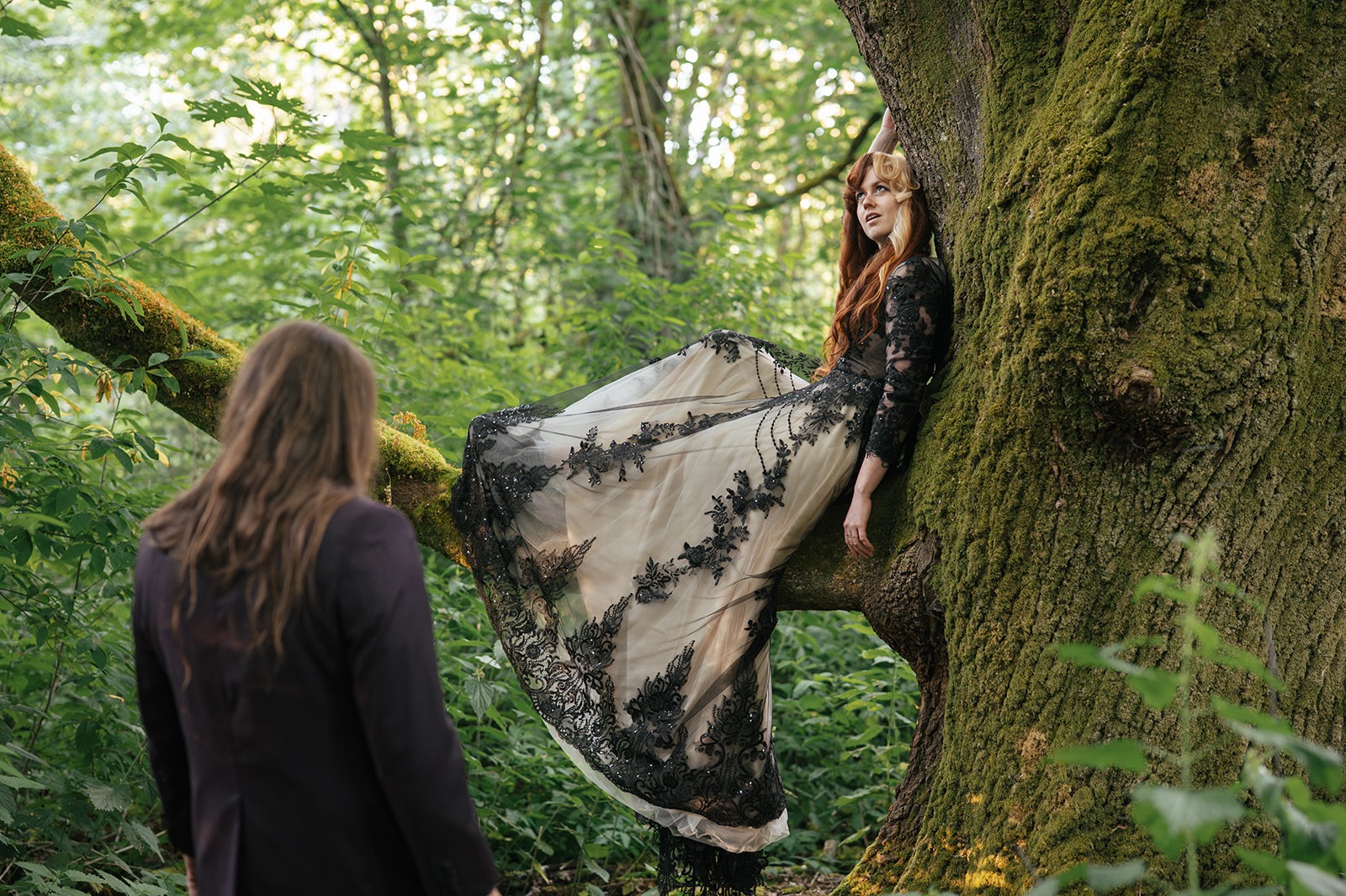 Bride sits in the crook of a moss-covered tree branch as the groom looks up at her
