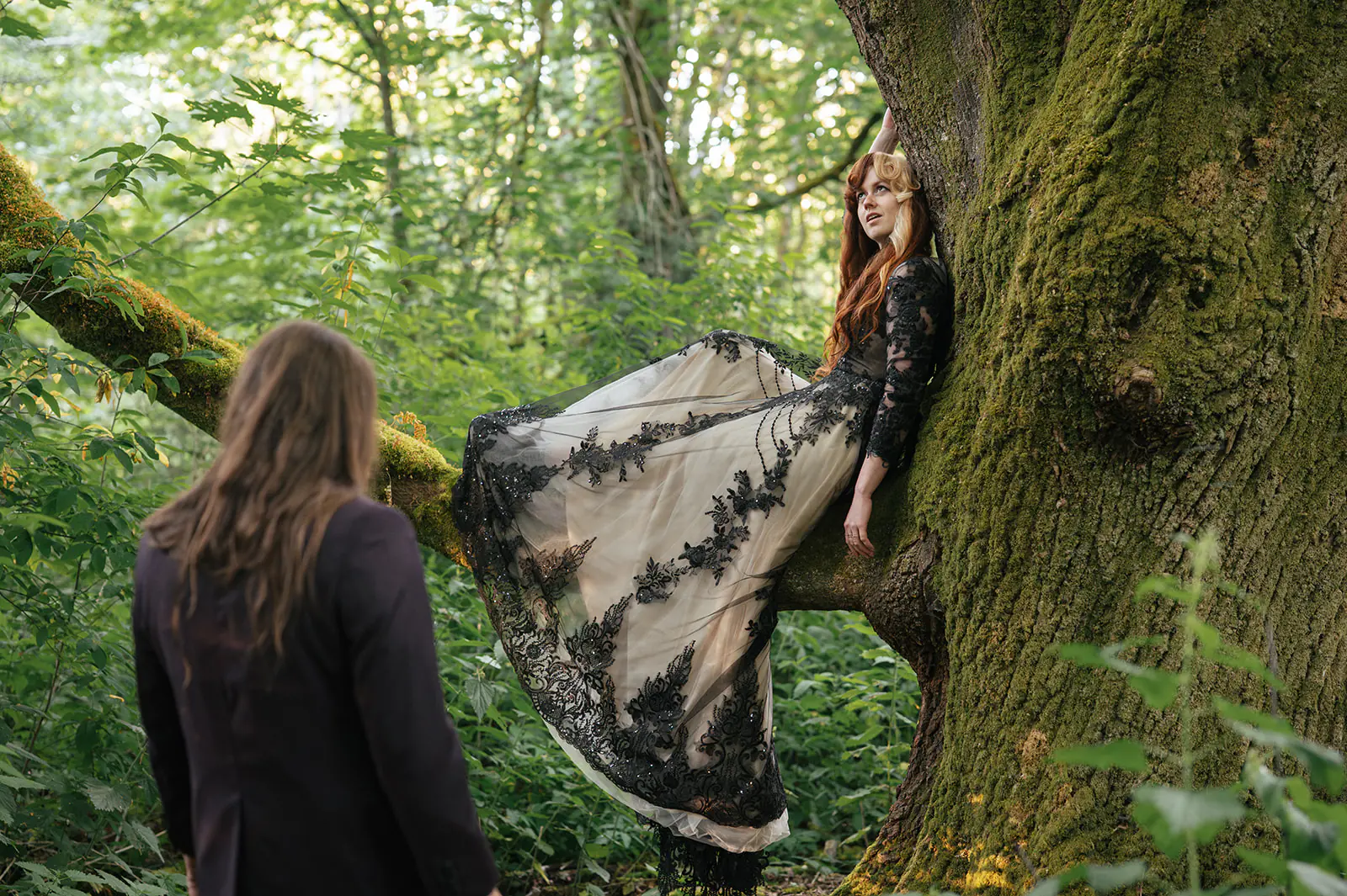 Bride sits in the crook of a moss-covered tree branch as the groom looks up at her