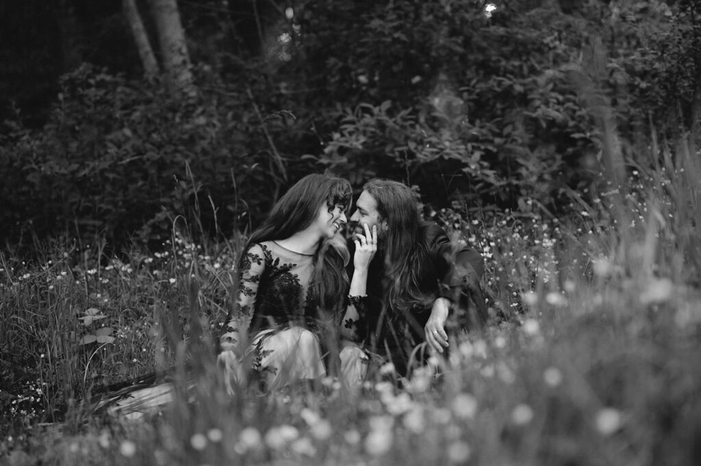 A black-and-white moody forest wedding portraits shot: couple sitting close in tall grass, foreheads touching