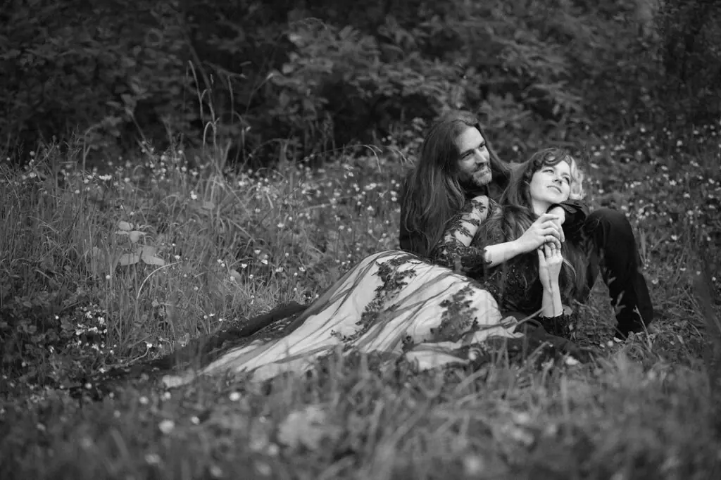 A black-and-white photo shows the couple sitting together in tall grass, groom offering the bride a bite