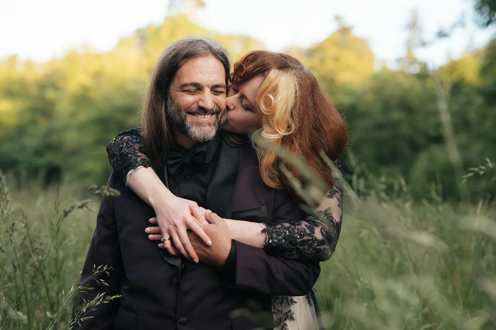 Couple shares a cheek kiss in a sunbeam-lit patch of forest during this moody forest wedding portraits session