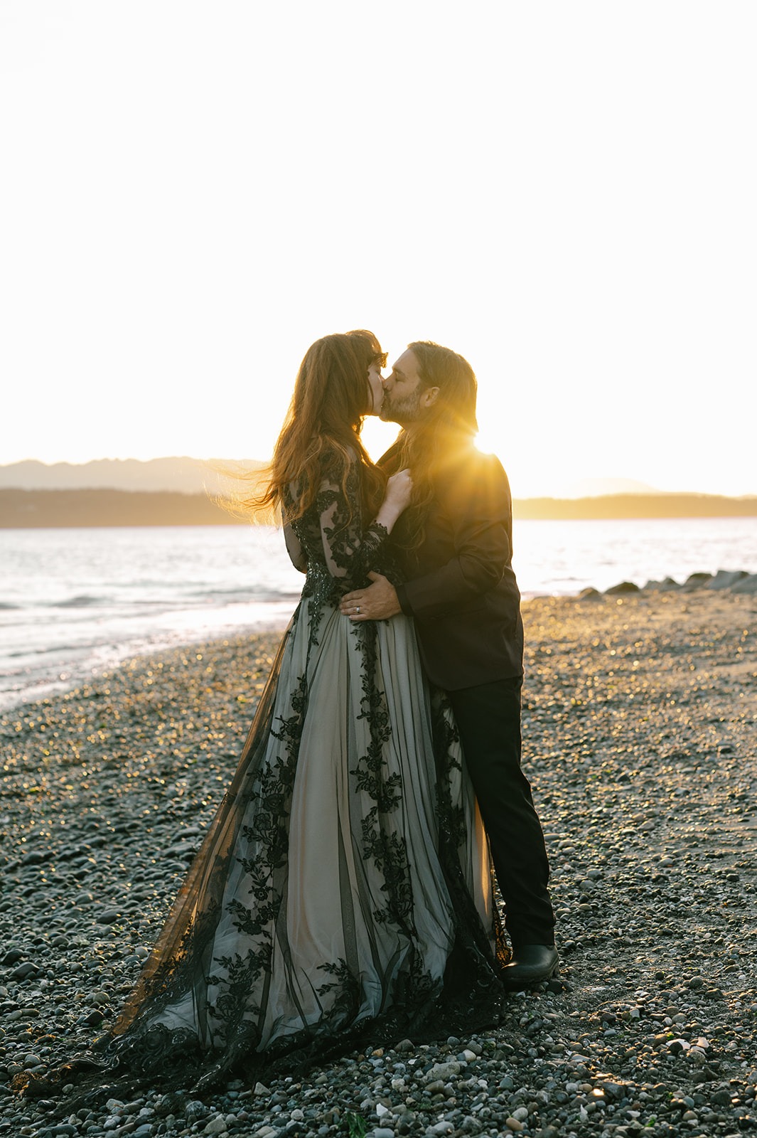 Couple kisses on the pebbled beach as the sun sets behind them