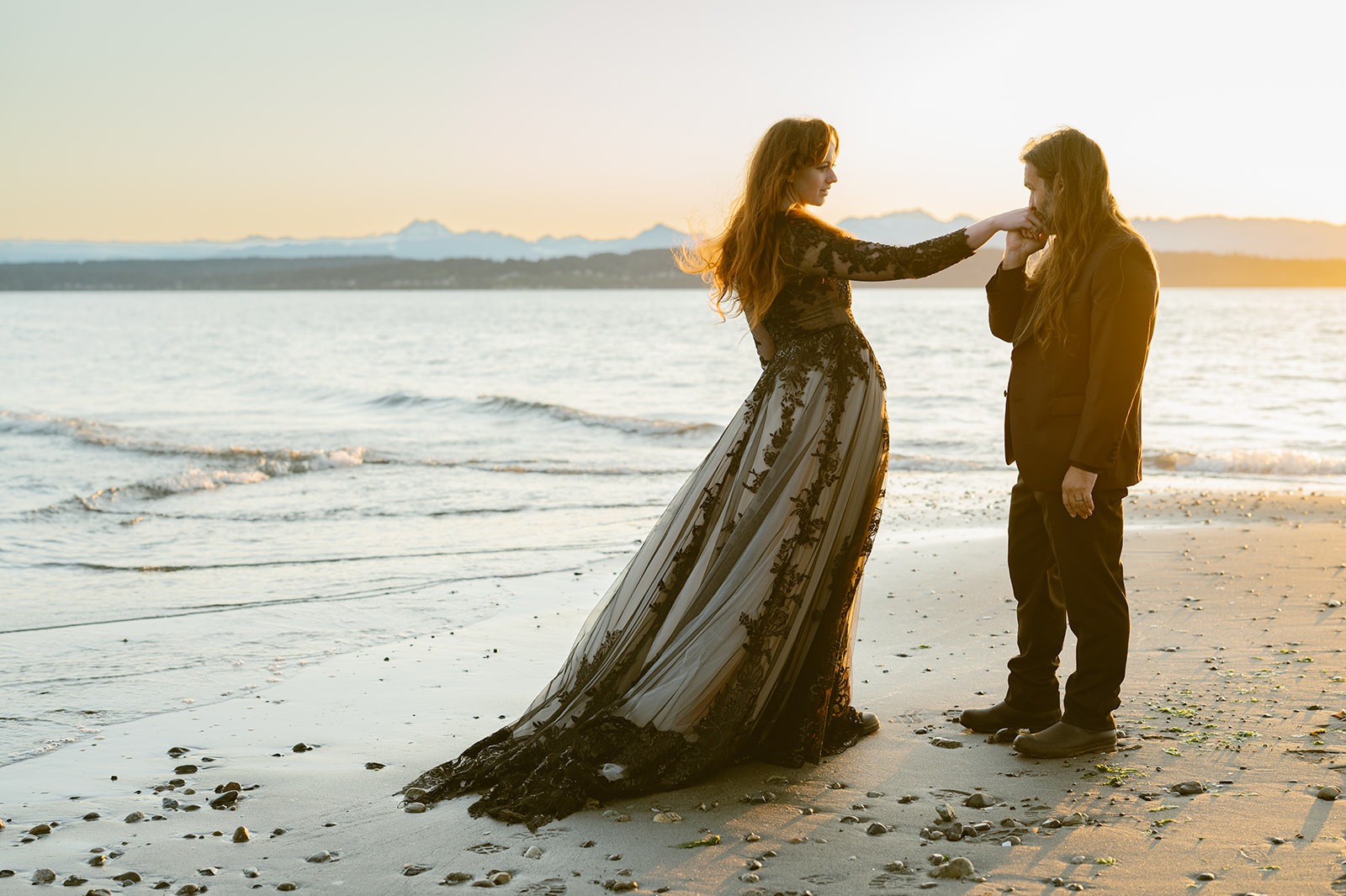 Bride reaches toward the groom's face as they stand together on the beach at sunset